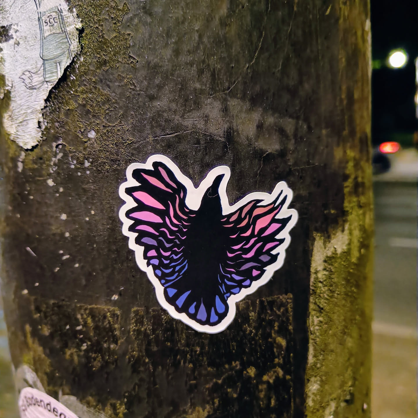 This sticker features a stylized bird with wings that look like they are made of feathers. The colors are a gradient from pink to purple to blue, and the bird itself is black. The image is striking, and the colors are eye-catching.