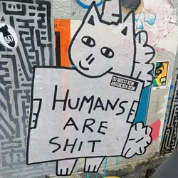 A sticker featuring a simple drawing of a cat holding a sign that says "Humans are shit." The cat is white with black outlines, and the sign is a dirty white color. There is another smaller sticker on the sign that says "DU MUSST KEIN ARSCHLOCH SEIN!" which translates from German to English as "YOU DON'T HAVE TO BE AN ASSHOLE!"