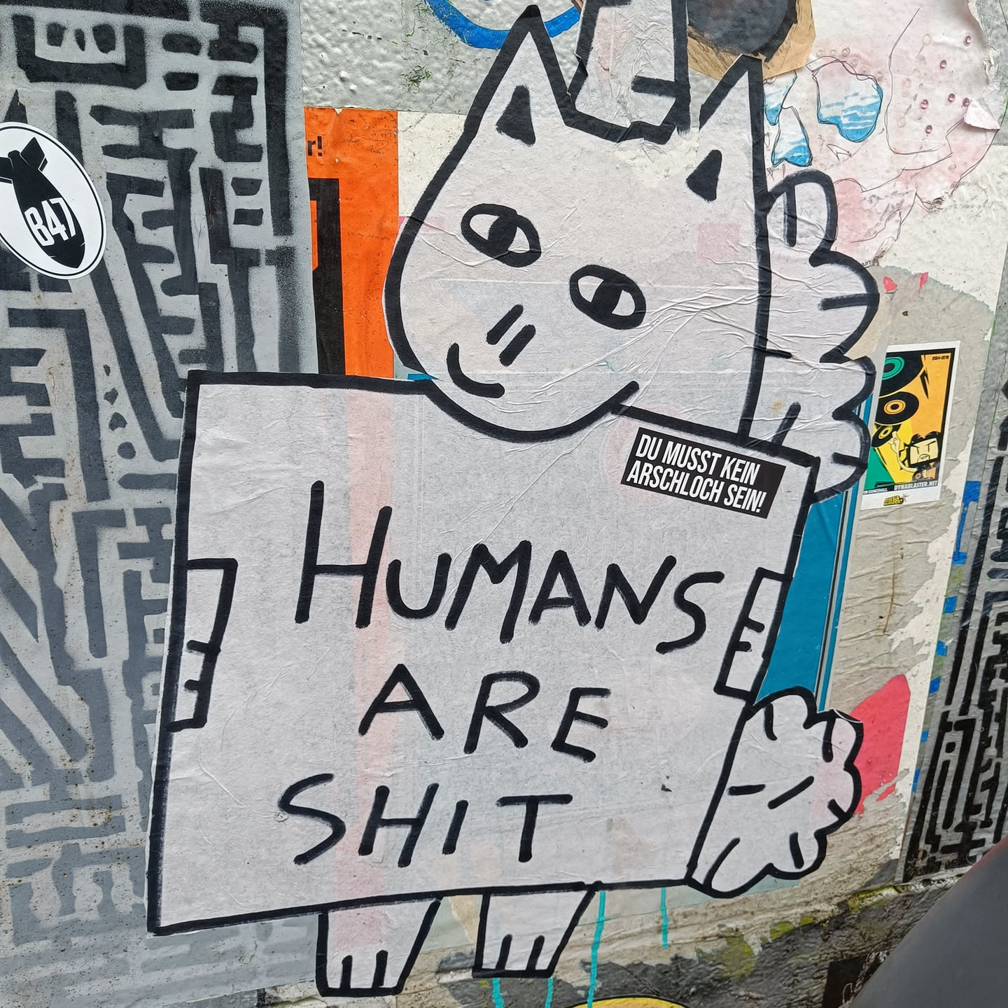 A sticker featuring a simple drawing of a cat holding a sign that says "Humans are shit." The cat is white with black outlines, and the sign is a dirty white color. There is another smaller sticker on the sign that says "DU MUSST KEIN ARSCHLOCH SEIN!" which translates from German to English as "YOU DON'T HAVE TO BE AN ASSHOLE!"