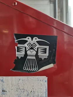 A black and white sticker featuring a stylized double bird design, possibly inspired by Native American art. The sticker is affixed to a red surface and shows some signs of wear.