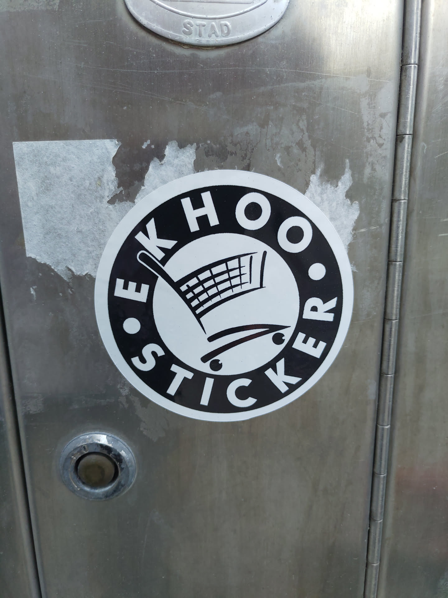 A black and white round sticker with the text "EKHOOS STICKER" arranged circularly around a simple shopping cart icon. The sticker is affixed to a metallic surface.