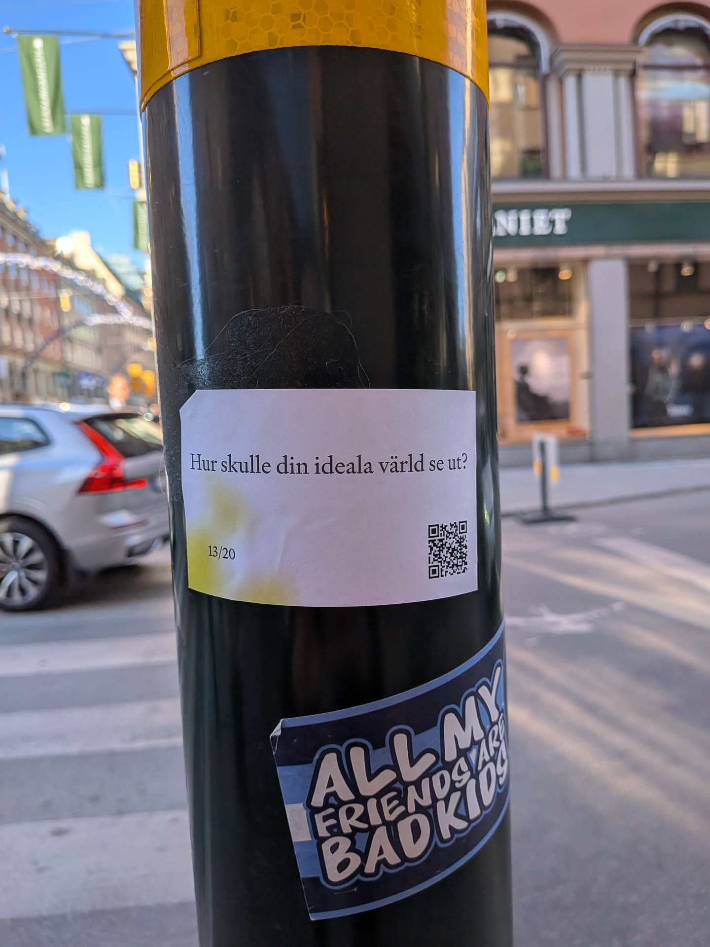 A rectangular white sticker adhered to a black pole. It features black text in Swedish asking 'Hur skulle din ideala värld se ut?' (What would your ideal world look like?) and the number '13/20'. To the right, a black and white QR code is prominently displayed. The bottom left corner of the sticker has a faint, yellowish-green gradient smudge.