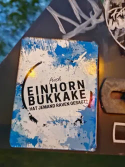 A sticker with a blue and white abstract background. The text on the sticker says "fresh Einhorn Bukkake" and "Hat jemand Raven gesagt?" in black. The text is in a circular shape, giving it a logo-like look. 