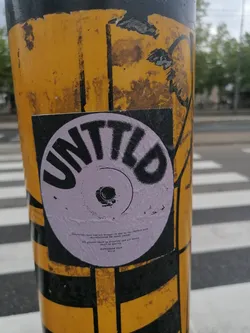 A circular sticker, primarily light purple with bold black text, is prominently displayed on a yellow and black striped pole. The main text, 'UNTITLD', is curved in an arc around the top, mimicking a vinyl record's label. In the center, there's a black dot within a larger circle, reinforcing the record theme. Below this, smaller black text provides more details, including: 'Immersion, bass and art brought to you by the UNTTLD team', 'manufactured for novel sounds', 'All grooves shall be grooving and all moves shall be moving', and 'ROTTERDAM 2024 RP110'. The pole itself shows signs of wear with peeling paint, and a blurred pedestrian crossing is visible in the background.