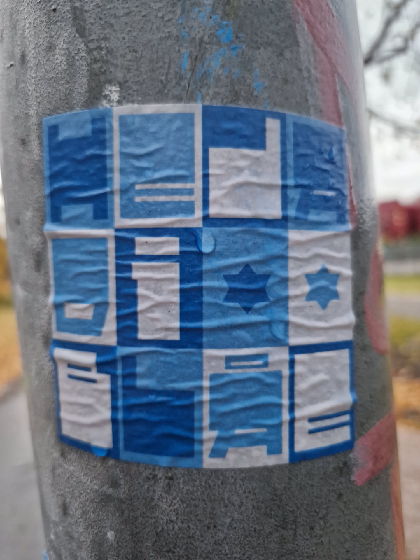 A sticker with a repeating geometric pattern in shades of blue and white.  The design includes simple shapes and symbols. The sticker is affixed to a metal pole outdoors and shows signs of weathering.