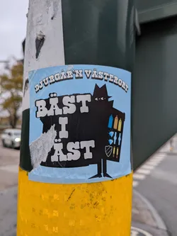 The sticker is predominantly light blue with a black silhouette of a figure wearing a fedora and trench coat. The figure appears to be holding a basket or bag filled with various items. Visible within the silhouette are three spray paint cans (blue, gold, yellow), two diamond shapes, a shield emblem, and two more spray paint cans (red and yellow). The text 'DJURGÅR'N VÄSTERORT' is arched across the top in white, outlined in black. Below this, in large, bold white letters with a black outline, is the text 'BÄST I ÄST'. The sticker shows signs of wear and tear, with some peeling and creasing, particularly on the left side.