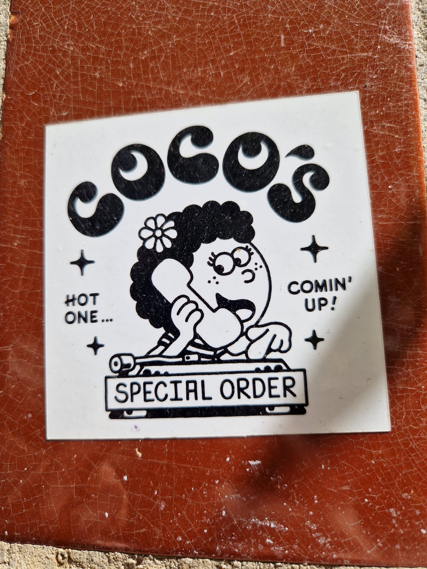 A sticker featuring a cartoon illustration of a girl behind a DJ mixer with the text "Coco's" at the top, "Special Order" at the bottom and "Hot one... Comin' up!" on either side. The style is reminiscent of 70s-80s cartoons.