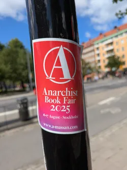 A sticker on a black pole promotes the Anarchist Book Fair 2025. The sticker has a red background that fades into pink, with a white anarchy symbol at the top. The text announces the event dates as 16-17 August and provides the website www.a-massan.com.