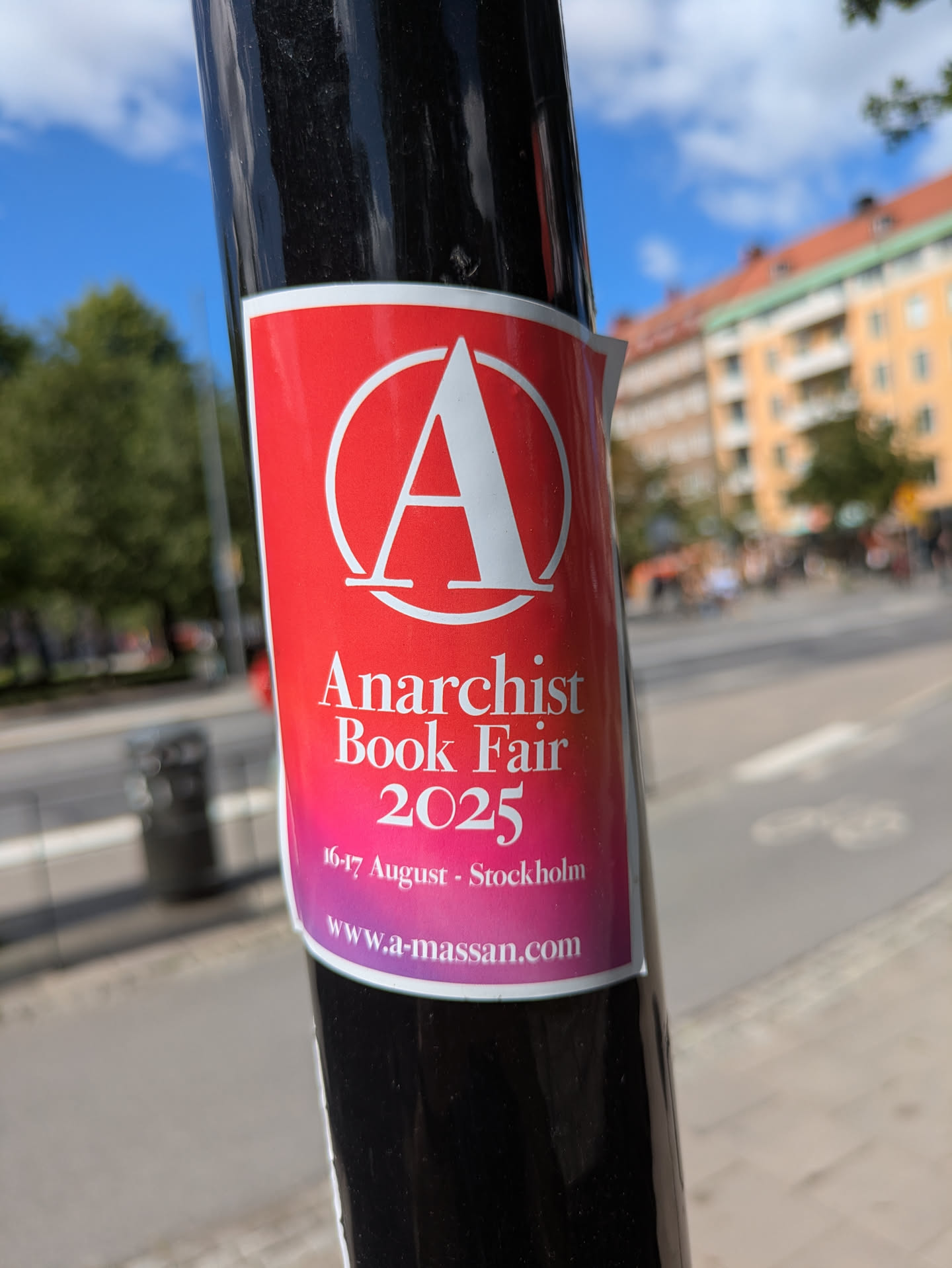 A sticker on a black pole promotes the Anarchist Book Fair 2025. The sticker has a red background that fades into pink, with a white anarchy symbol at the top. The text announces the event dates as 16-17 August and provides the website www.a-massan.com.