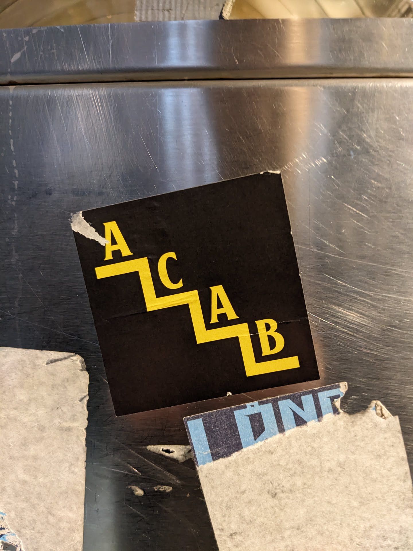 A black and yellow sticker with the acronym ACAB arranged as steps.  The sticker is affixed to a metallic surface.