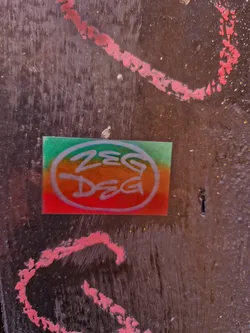 A small, circular sticker with the tag 'ZEG DEG' written in a stylized font. The sticker has a gradient background of green, orange and red. 