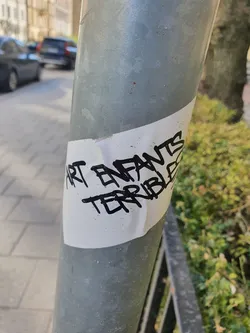 A white sticker with black marker writing on a grey metal pole. The text says "Art Enfants Terribles".