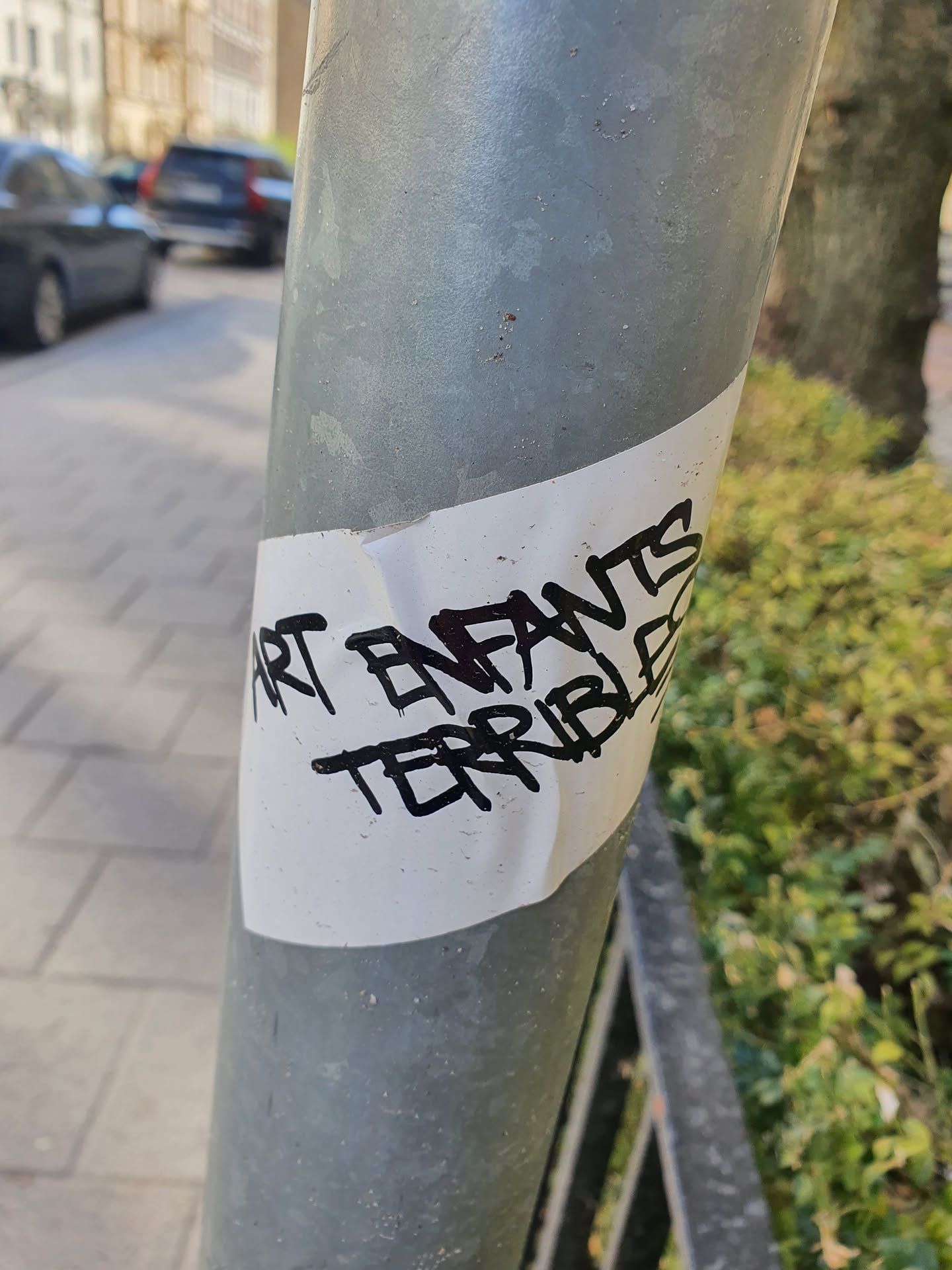 A white sticker with black marker writing on a grey metal pole. The text says "Art Enfants Terribles".