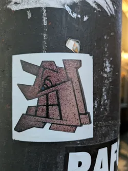The sticker features a stylized, abstract 'A' shape. It is rendered in shades of brown and rust, with thick black outlines defining its geometric segments. The texture appears somewhat rough and mottled, giving it an aged or industrial appearance. A small blue dot is visible within the central negative space of the letterform. The sticker is white-bordered and centered on a dark, textured cylindrical surface.