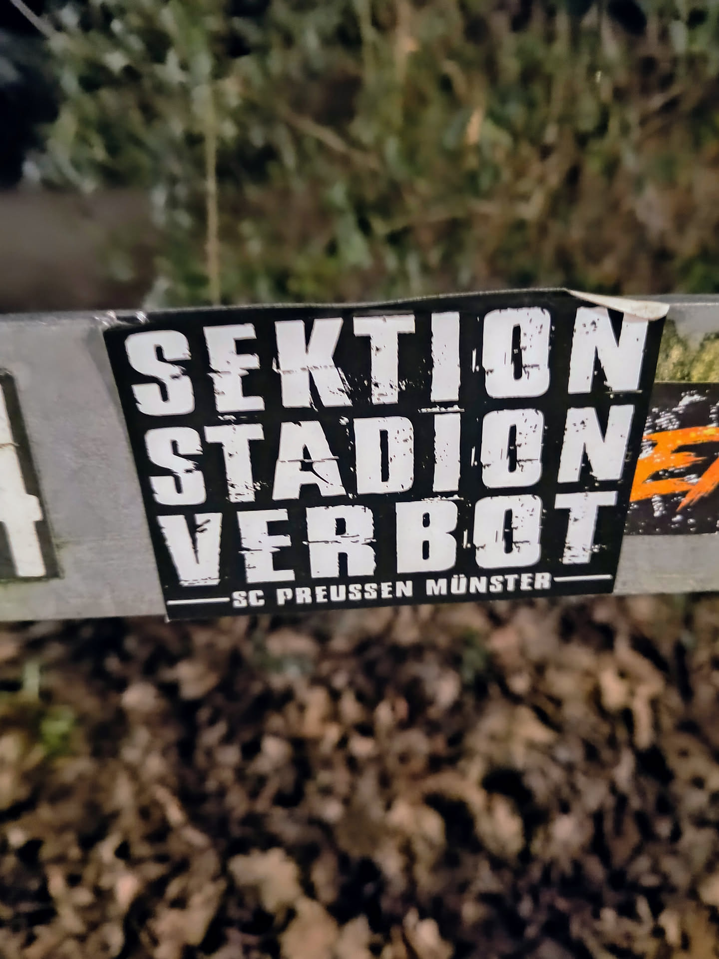 A black and white sticker with the text "SEKTIO
STADION
VERBOT" and the smaller text "-SC Preussen Münster- at the bottom. The sticker is attached to a surface outdoors.