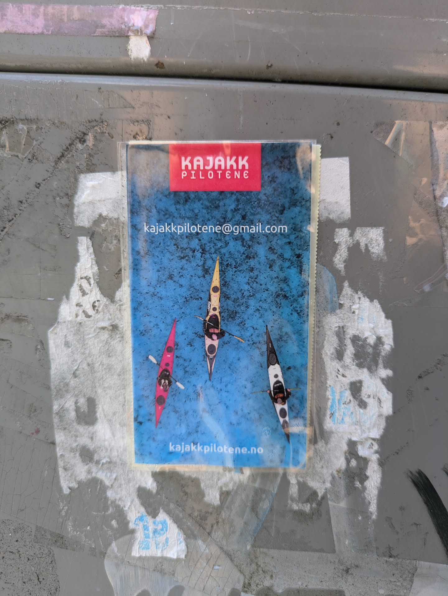 The sticker prominently displays an aerial shot of three individuals in kayaks (one pink, one yellow, and one black and white) paddling on a vibrant blue body of water. At the top, there is a red rectangular banner with the white text 'KAJAKK PILOTENE'. Below this, in white text, is the email address 'kajakkpilotene@gmail.com'. At the very bottom of the sticker, also in white text, is the website address 'kajakkpilotene.no'. The sticker is adhered to a weathered, grey surface.