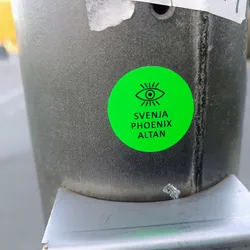 Close-up of a round, bright green sticker affixed to a gray metal pole. The sticker features a simple line drawing of an eye with radiating lines, and the text "SVENJA PHOENIX ALTAN" is printed below the eye symbol in black font. The overall style is minimalist and modern.