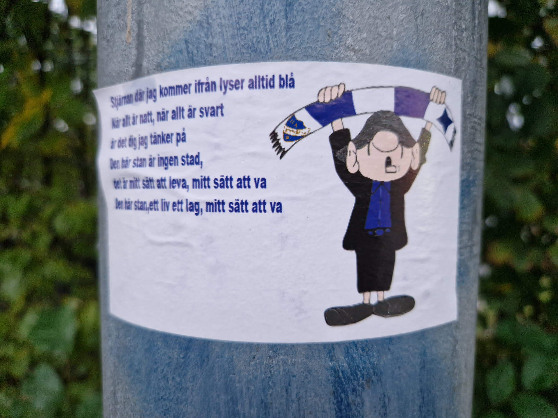 The sticker shows a simple cartoon drawing of a person holding up a blue and white scarf.  A poem in Swedish is printed on the sticker. The poem seems to be about the person's hometown and way of life.