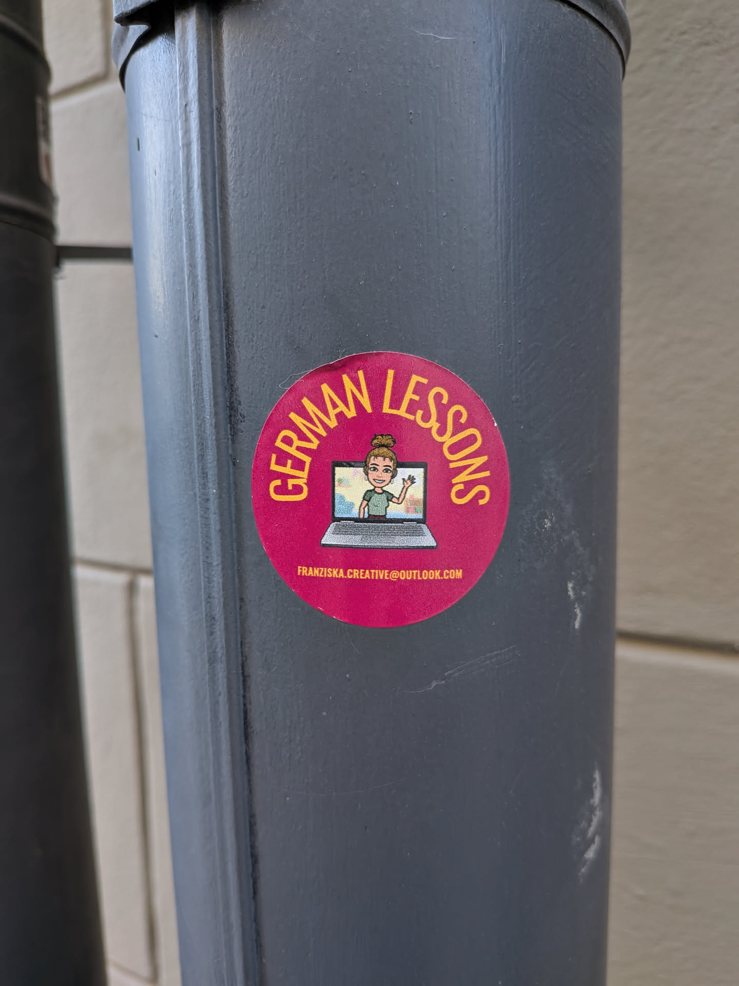 A circular sticker with a vibrant magenta background. The top half features the words 'GERMAN LESSONS' in yellow capital letters, curving along the edge. In the center, there's an illustration of a silver laptop with a screen displaying a cartoon avatar of a woman. The avatar has light brown hair in a bun, green eyes, and is wearing a green top, waving with her right hand. Below the laptop, in smaller white text, an email address is visible.