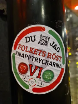 A sticker with a QR code, thumbs up/down and the text "DU FOLKETS RÖST KNAPPTRYCKARNA JAG VI".  It's designed to look like a button with green and red sections, indicating a choice.