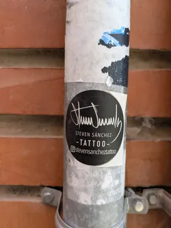 This black, round sticker, attached to a weathered metal pole against a brick wall, features the logo and contact information for Steven Sánchez Tattoo. The design includes a stylized signature above the text "-STEVEN SÁNCHEZ-" and "-TATTOO-". The Instagram handle @stevensancheztattoo is clearly displayed at the bottom.