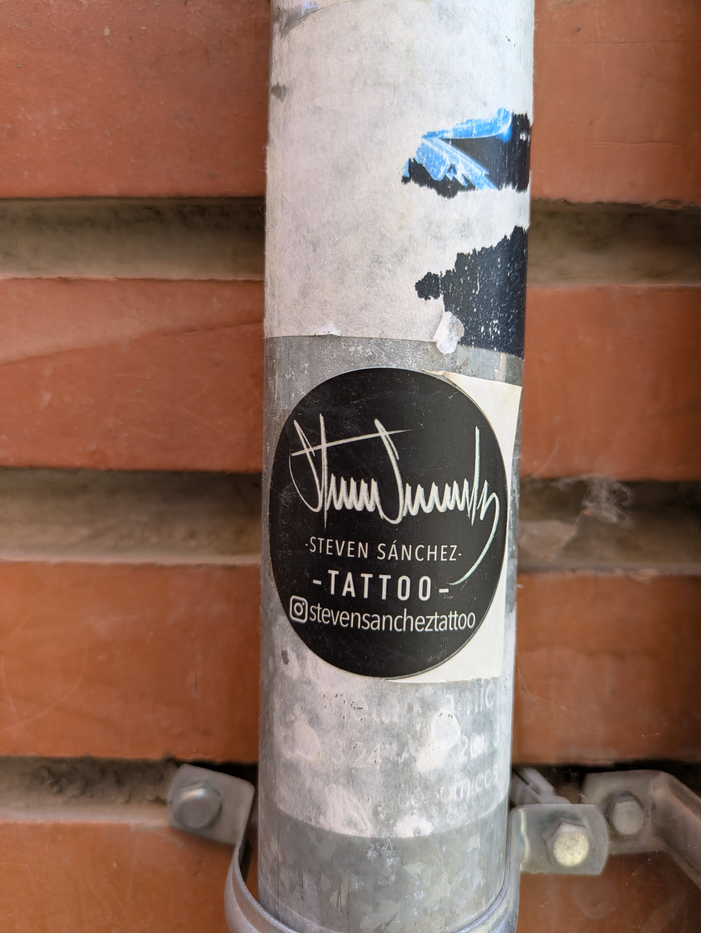 This black, round sticker, attached to a weathered metal pole against a brick wall, features the logo and contact information for Steven Sánchez Tattoo. The design includes a stylized signature above the text "-STEVEN SÁNCHEZ-" and "-TATTOO-". The Instagram handle @stevensancheztattoo is clearly displayed at the bottom.