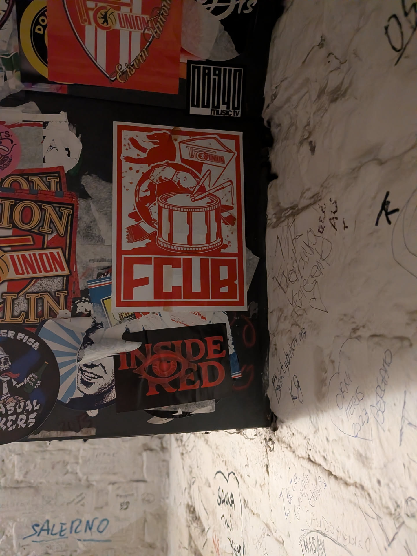 A vertically oriented rectangular sticker printed in red monochrome on a white background. The illustration displays classic football supporter paraphernalia: a marching snare drum with drumsticks, a football, a supporter scarf, a waving fan flag bearing the '1. FC UNION' logo, and a silhouette of the Berlin bear in the upper left corner. Below the central illustration is the large, bold abbreviation 'FCUB' enclosed in a rectangular border.