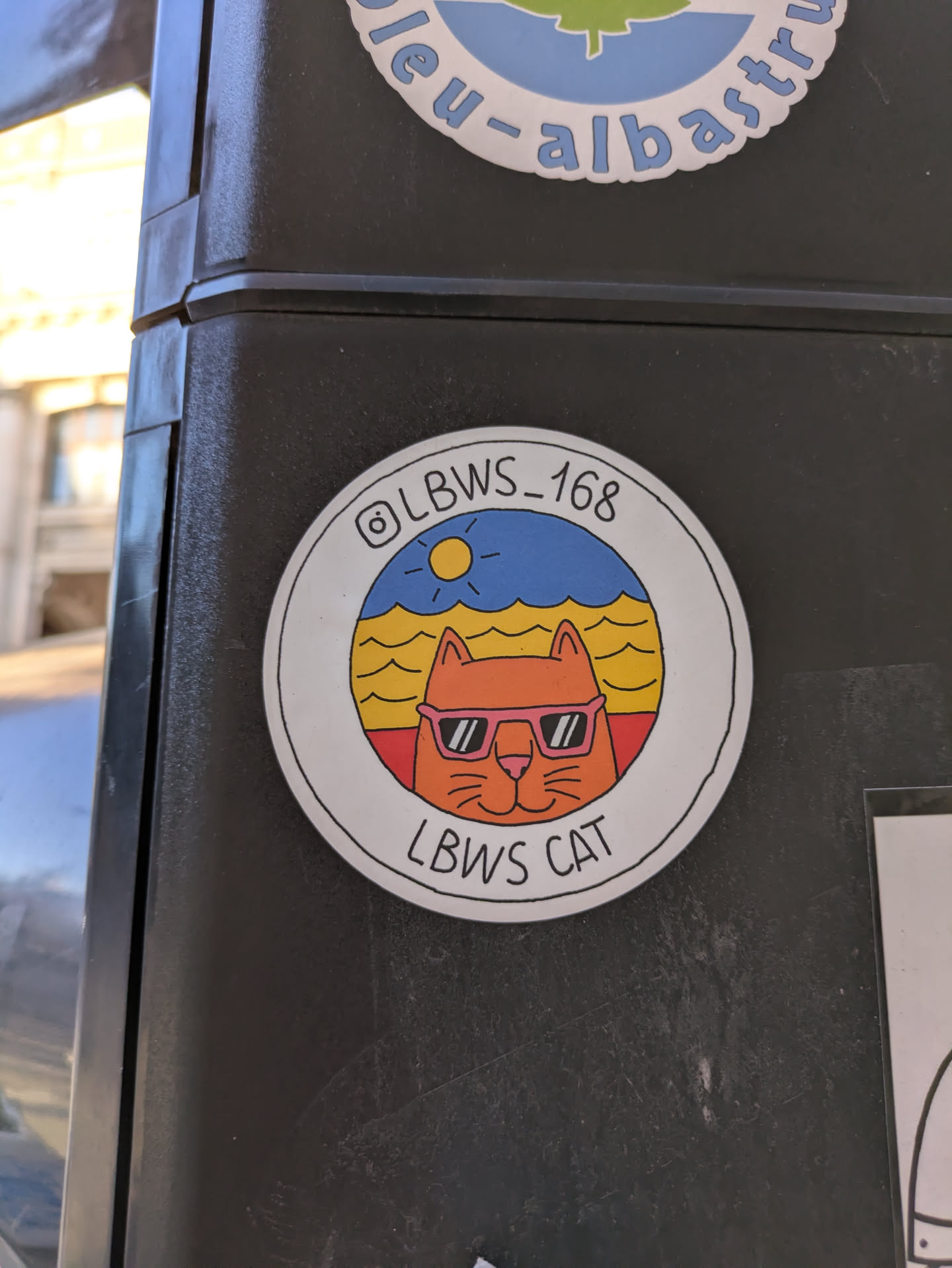 A round sticker featuring a cartoon cat with sunglasses against a backdrop of an ocean scene with a sun. The text reads "LBWS_168" above the cat and "LBWS CAT" below.