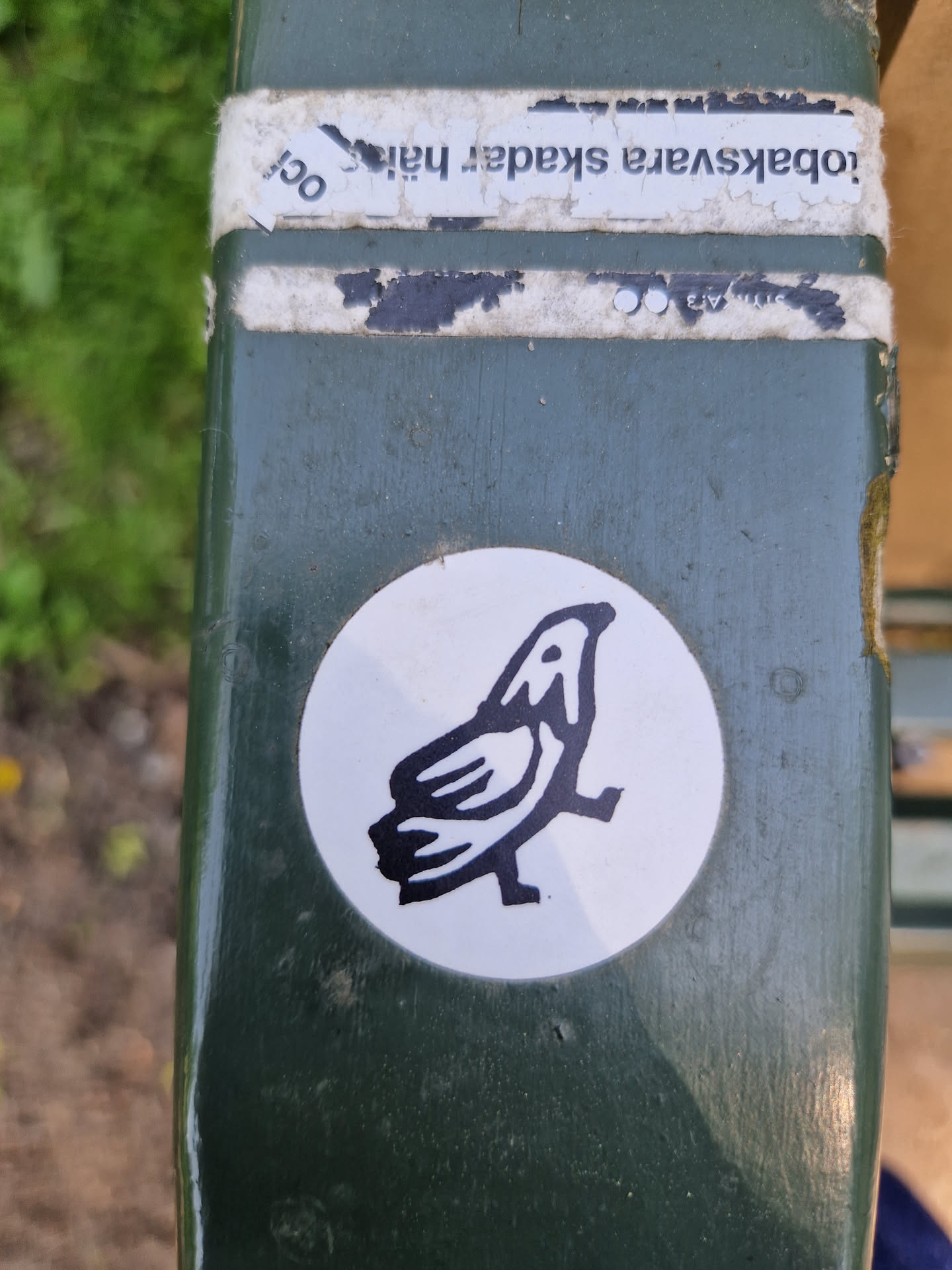 A round white sticker with a black illustration of a bird. The sticker is attached to a dark-green surface. Some old, damaged stickers are visible above it. 