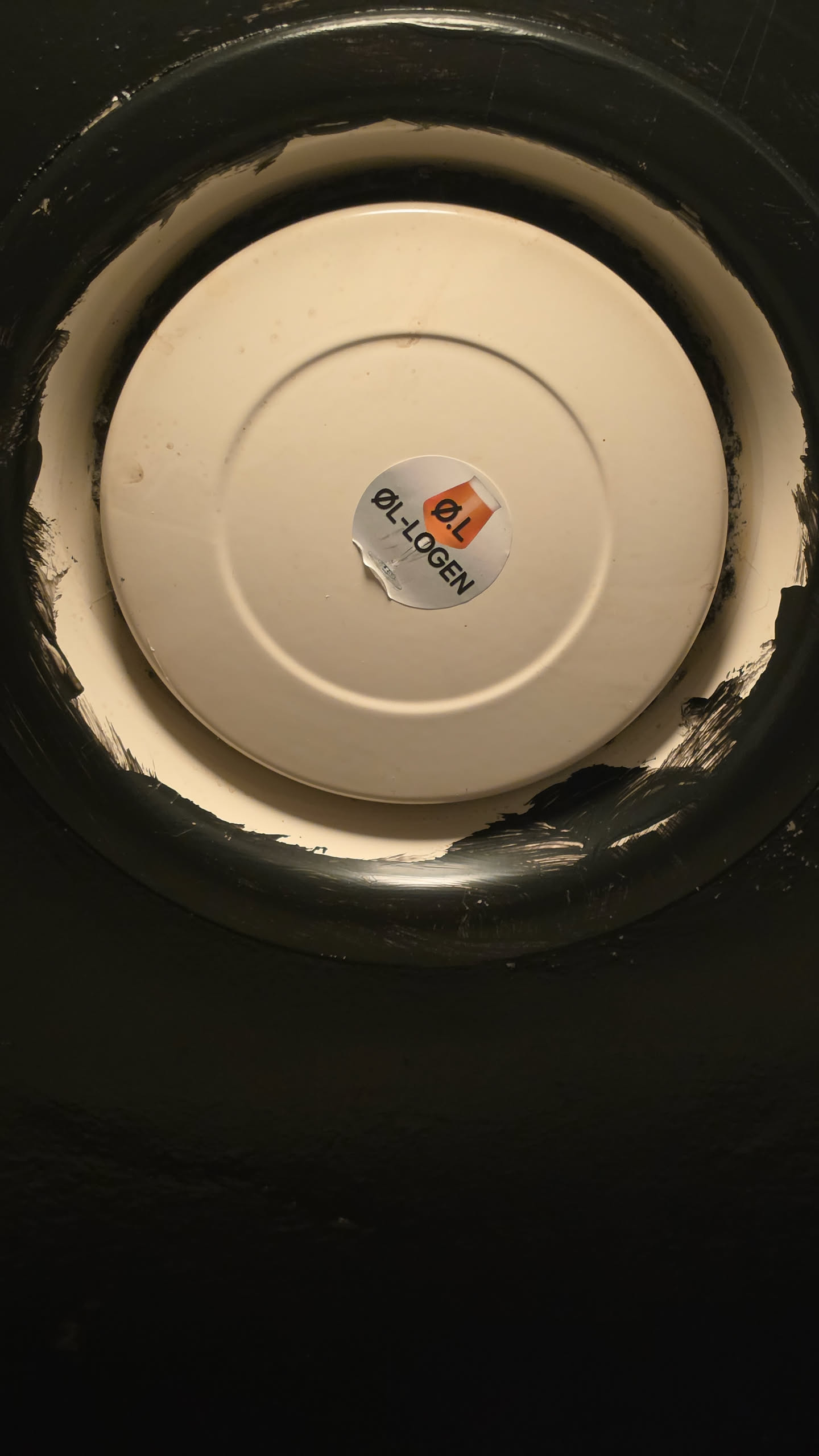 A circular sticker with a white background positioned in the center of an off-white circular surface. It features a graphic of a beer glass filled with amber-colored liquid and topped with white foam, overlaid with the bold text 'Ø.L'. Below the icon, the text 'ØL-LOGEN' curves in capitalized black sans-serif font. The lower left edge of the sticker is slightly crumpled and peeling.