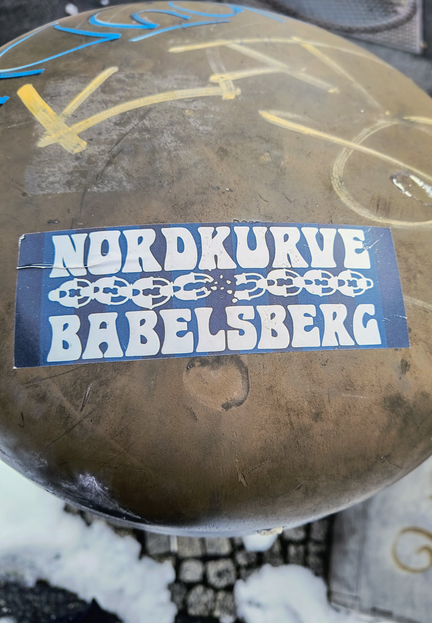 A sticker featuring the text "Nordkurve Babelsberg" in a retro font, with a graphic of a broken chain underneath. The sticker is affixed to a weathered surface.