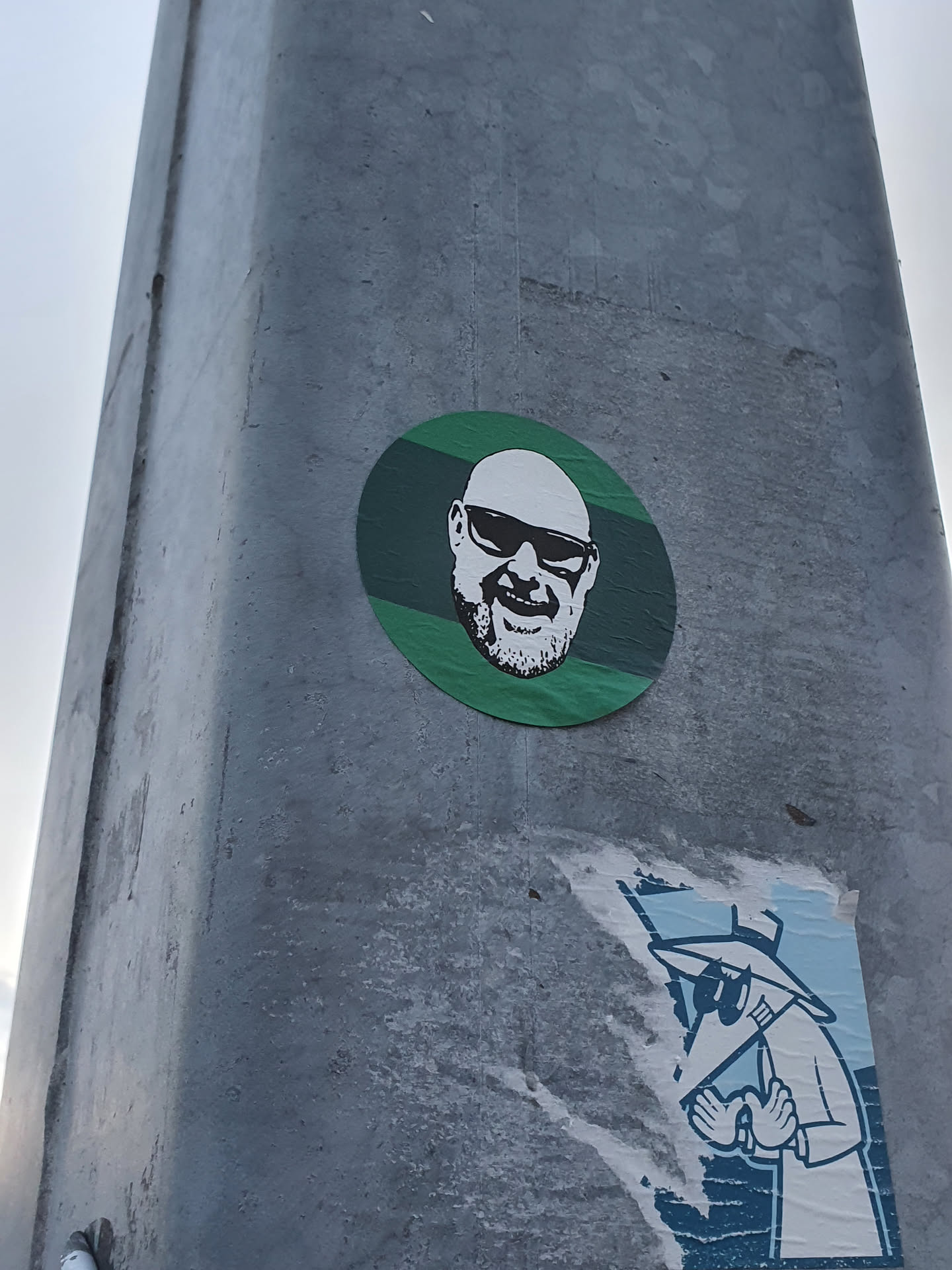A round sticker featuring a portrait of a bald man wearing sunglasses. The portrait is in black and white, and the background is green with horizontal stripes.  There's a second, partially torn sticker below it featuring a figure in a hat and coat.