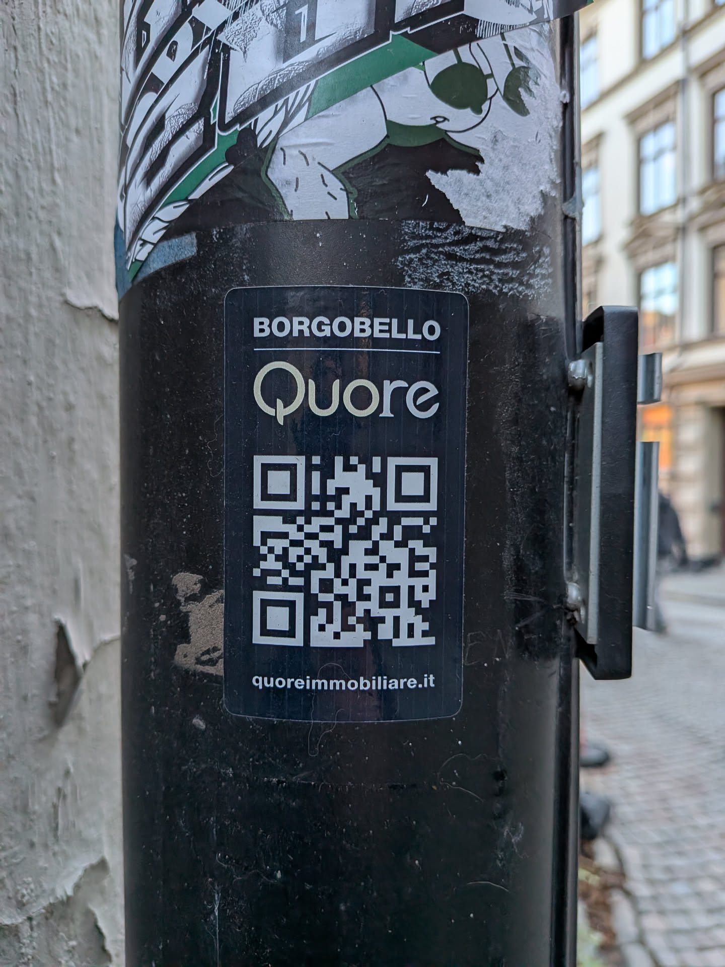 A rectangular black sticker featuring white text and a yellow-gold logo. At the top, 'BORGOBELLO' is written in white uppercase letters. Below it, the word 'Quore' is prominently displayed in a stylized yellow-gold font, with the 'Q' forming a heart-like shape. A large, white QR code is centered below the logo. At the very bottom, 'quoreimmobiliare.it' is written in smaller white text. The sticker is applied to a dark pole and shows some minor dust and wear, indicating it's a real-world application.