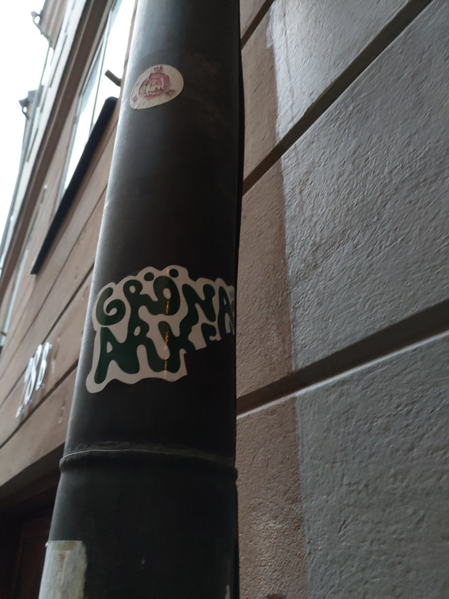 A green and white sticker with the words "GRÖNA ARKET" in a stylized font. The sticker is affixed to a dark gray metal pole on the side of a building.