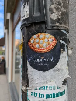 A round sticker on a pole shows an image of a pizza at the top and the word "Suprema" and the phrase "Sinulle tehty" at the bottom. The background behind the text is marble.