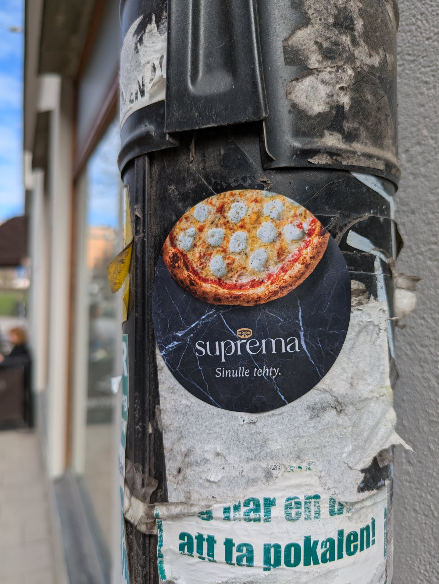 A round sticker on a pole shows an image of a pizza at the top and the word "Suprema" and the phrase "Sinulle tehty" at the bottom. The background behind the text is marble.