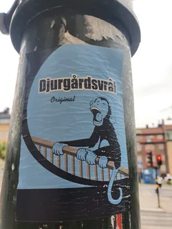 A sticker featuring a cartoon monkey sitting on a railing, with Djurgårdsvarå Original written on it. The sticker is light blue with a black outline and the monkey is dark blue. The background is a light blue circle. The sticker is affixed to a dark green pole.