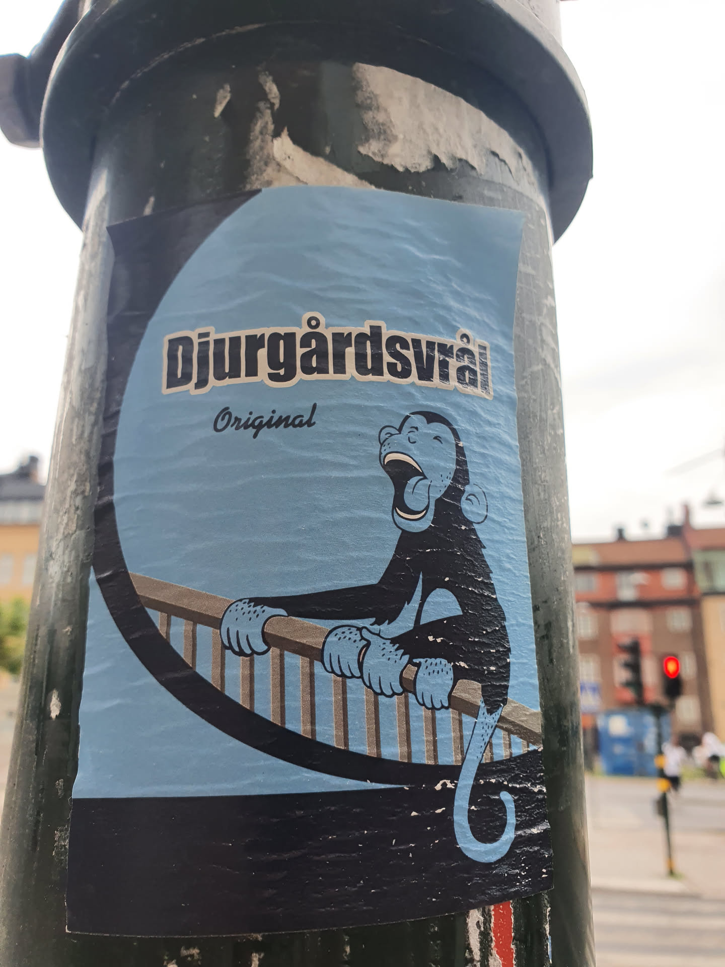 A sticker featuring a cartoon monkey sitting on a railing, with Djurgårdsvarå Original written on it. The sticker is light blue with a black outline and the monkey is dark blue. The background is a light blue circle. The sticker is affixed to a dark green pole.