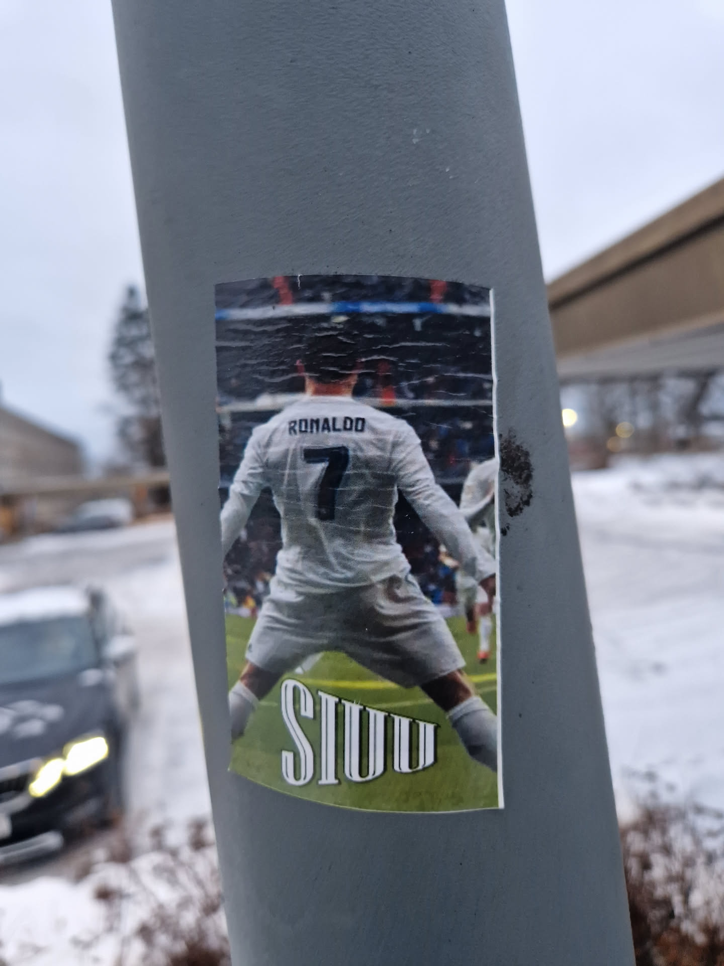A sticker featuring Cristiano Ronaldo with his back turned, celebrating a goal with his signature 'Siuu' pose. The sticker is attached to a grey pole in a snowy setting.