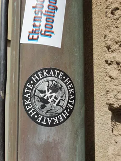 Round black and white sticker with the name HEKATE repeating around the border. The center features a stylized image that seems to be related to the goddess Hecate. 