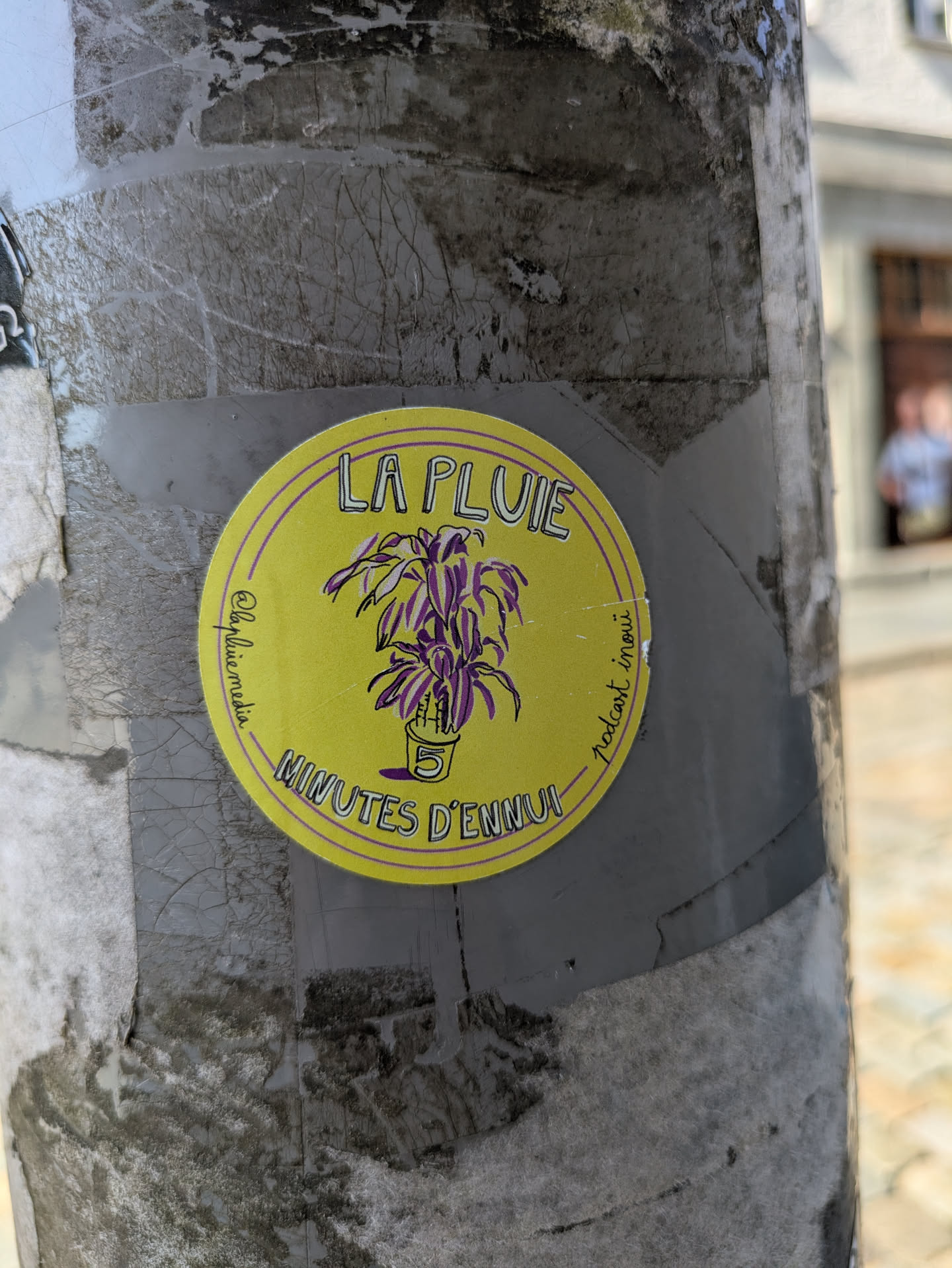 A vibrant yellow circular sticker adhered to a weathered gray pole. The sticker features an illustration of a potted plant with purple and green leaves. The pot has a white '5' on it. Around the top edge of the circle, the text 'LA PLUIE' is visible, and along the bottom edge, 'MINUTES D'ENNUI' is written. On the left side, vertically, it reads '@lapluie.media' and on the right side, vertically, 'podcast inoui'. The sticker has a thin purple outline.