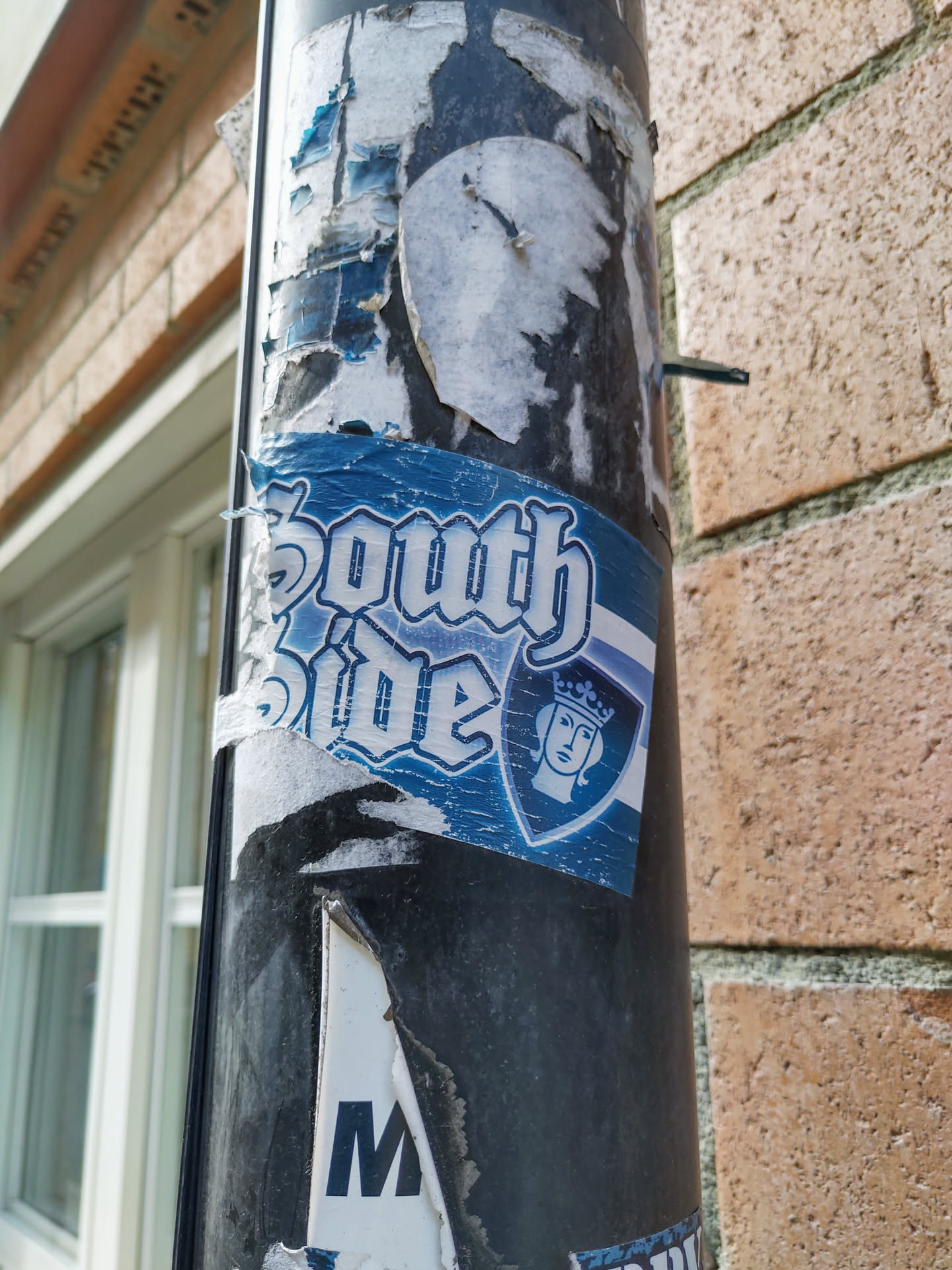 A blue and white sticker featuring the words "South Side" in a stylized font and a crest with a woman's face wearing a crown.  The sticker shows some wear and tear, suggesting it's been up for a while. The background is a dark pole covered in other old stickers.