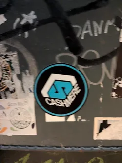 Street sticker A circular sticker set against a dark, graffitied surface. It features a black background bordered by a vibrant light-blue ring. In the center is a stylized geometric emblem comprising an angular white outer shape and a bright cyan interlocking letter 'S' motif. Underneath the emblem, the word 'CASHMERE' is printed in bold white uppercase lettering following the curve of the lower edge.