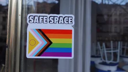 The sticker is rectangular with rounded corners, featuring a clean white border. At the top, in bold, sans-serif black letters with a white outline and a subtle drop shadow, is the text "SAFE SPACE". Below the text is a depiction of the Progress Pride Flag. This flag design incorporates the traditional six-stripe rainbow flag (red, orange, yellow, green, blue, purple) on the right side. On the left, a chevron points to the right, composed of five stripes: light blue, pink, and white (representing the transgender flag), followed by brown and black stripes (representing marginalized people of color and those living with AIDS or lost to the disease). Within the yellow section of the chevron, there is a thin purple circle. The sticker is affixed to a reflective glass surface, with reflections of the surroundings visible.