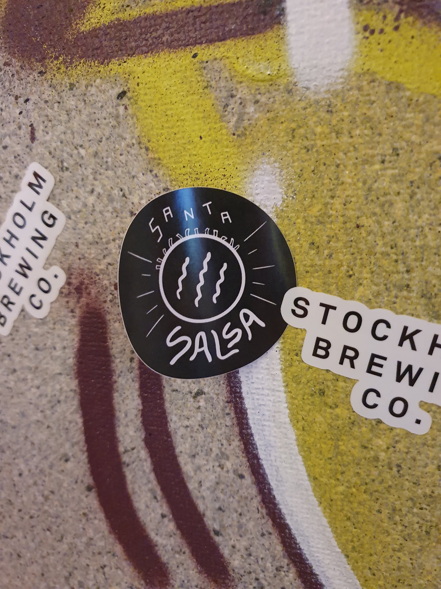 A black round sticker with the text 'SANTA SALSA' in a stylized font. It also features a simple graphic of three wavy lines inside a circle, which presumably represents beer.  The sticker appears to be placed on a wall with other stickers and graffiti.