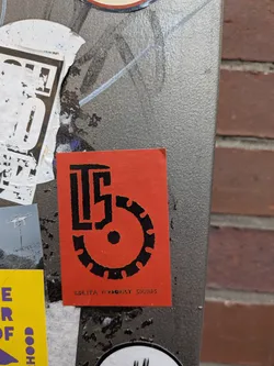 A vertical rectangular sticker in vivid red, printed with bold black ink. It features a stylized monogram emblem composed of the letters 'LTS' integrated with a notched circular gear shape. Below the graphic, the text 'LOLITA TERRORIST SOUNDS' is printed in a clean, small sans-serif typeface.