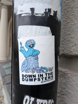 Street sticker A rectangular white sticker featuring a cartoon mashup of Cookie Monster and Oscar the Grouch. The blue furry monster emerges from a silver-blue trash can holding an opened tin can, with garbage such as banana peels, jars, cans, and an apple core scattered around the base. Below the graphic, bold black sans-serif typography reads 'DOWN IN THE DUMPSTERS', accompanied by the handle '@JAYVELCRO' in smaller print in the lower right corner.