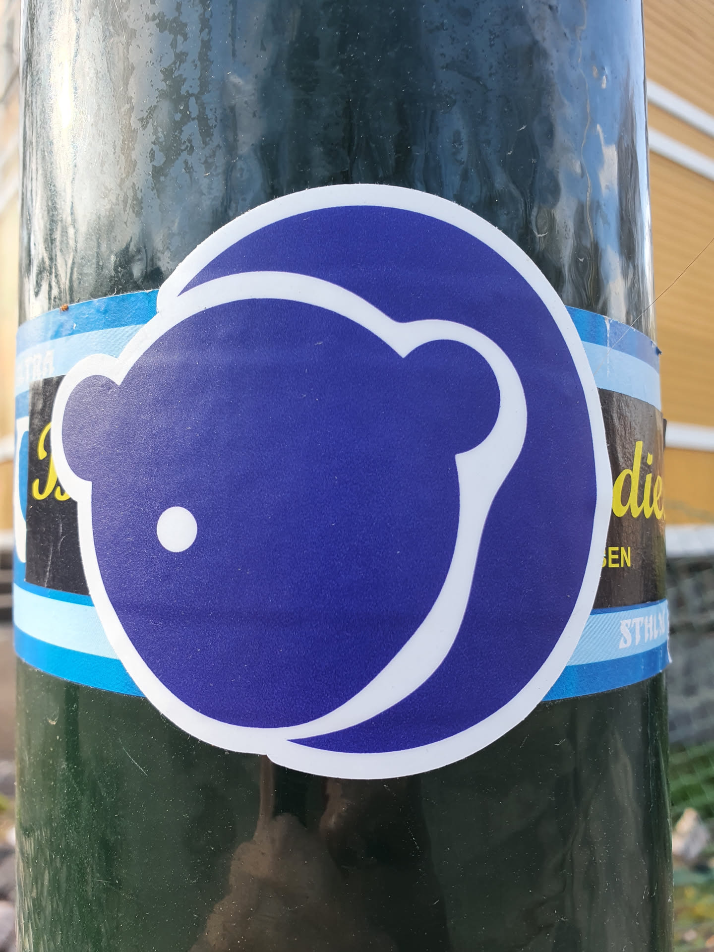 A round sticker featuring a stylized purple bear. The bear is simple, with a single dot for an eye and a slightly curved body.  The sticker has a white outline. It's affixed to a dark green pole.