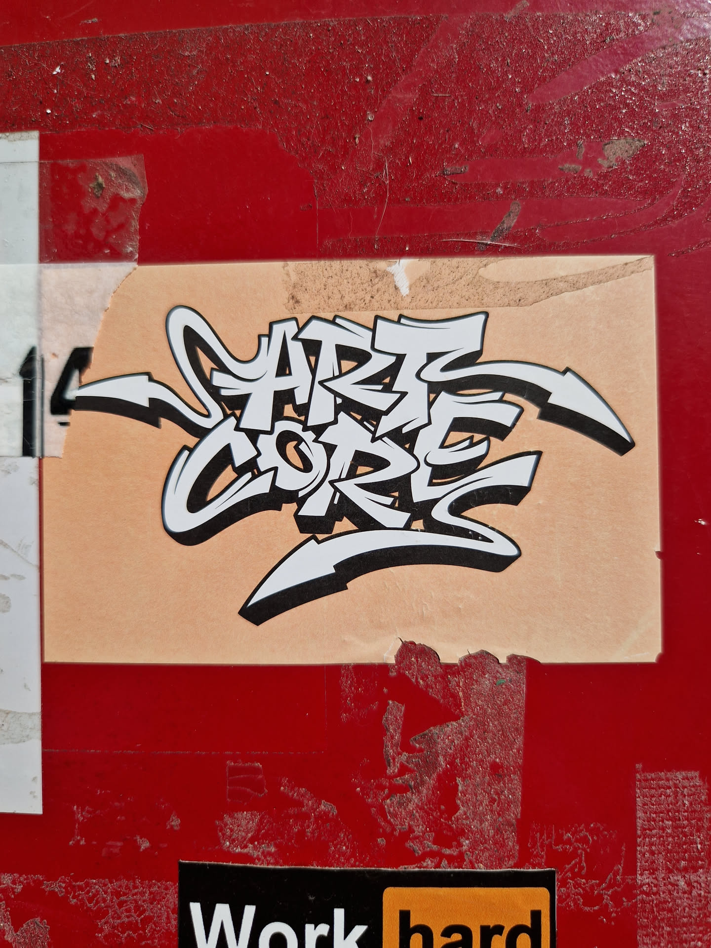 A beige sticker with black and white graffiti art that says \"Art Core\" in a stylized font. The sticker is attached to a red surface and shows some signs of wear.