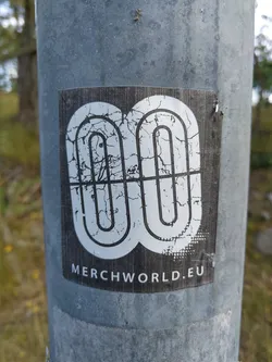 This sticker features a stylized logo resembling interconnected loops, presented in a distressed, cracked style.  The text 'MERCHWORLD.EU' is visible at the bottom.  The sticker appears to be affixed to a metal pole outdoors.